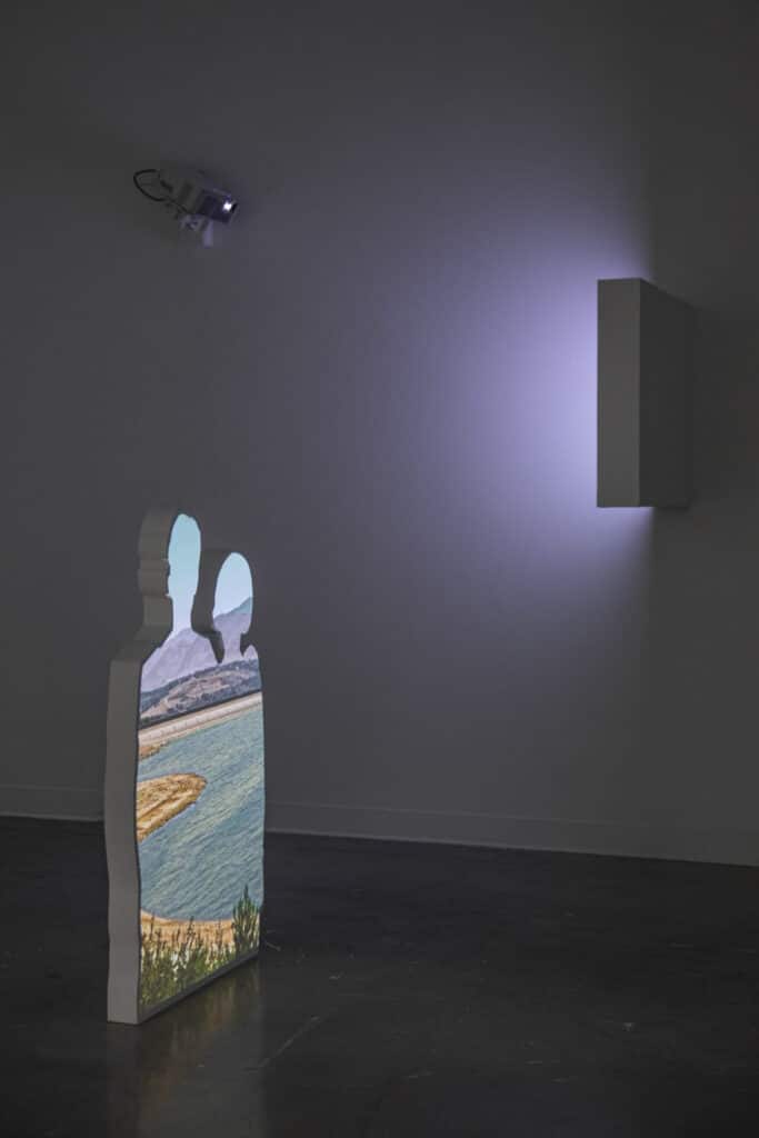 A projected image of a landscape with mountains and water appears on a cutout silhouette of two figures standing in a dark gallery space. A soft purple light glows on the wall beside them, creating a contemplative, cinematic atmosphere.