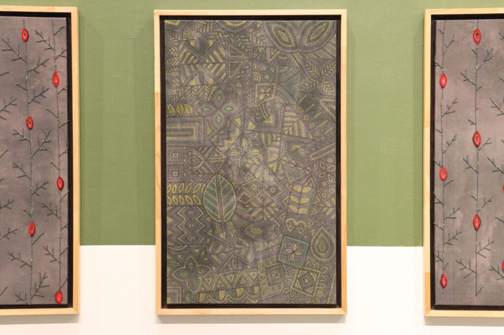 A textile artwork featuring intricate embroidered geometric and organic patterns in muted greens and yellows hangs in a wooden frame. The piece is flanked by panels with red embroidered motifs on gray fabric, displayed against a green and white wall.