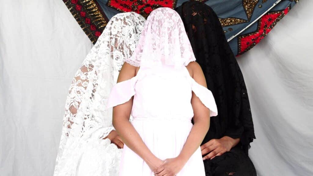 Three figures pose against a draped fabric backdrop, their faces covered with lace veils—one in white, one in pink, and one in black. The central figure stands in a pale pink dress, framed by the seated figures on either side, creating a striking, symbolic composition.