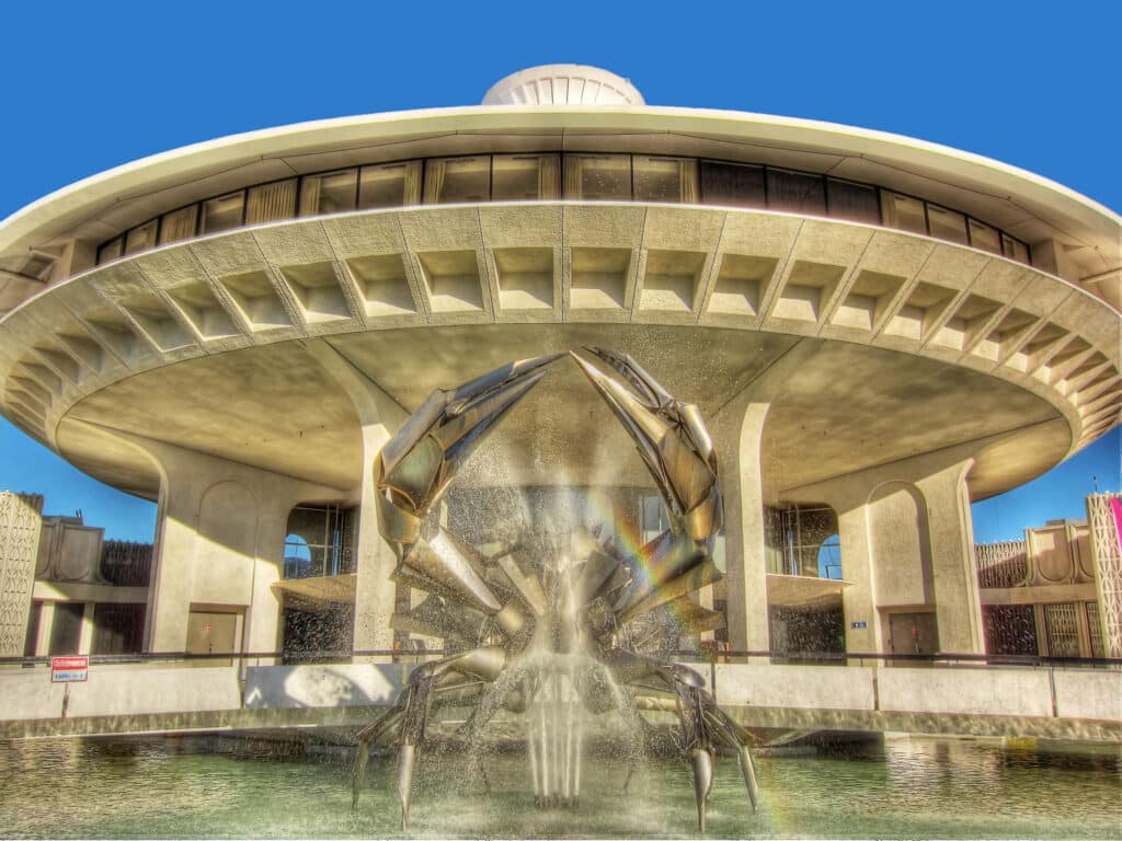 A metal crab sculpture sits in the centre of a fountain in front of a circular modernist building.