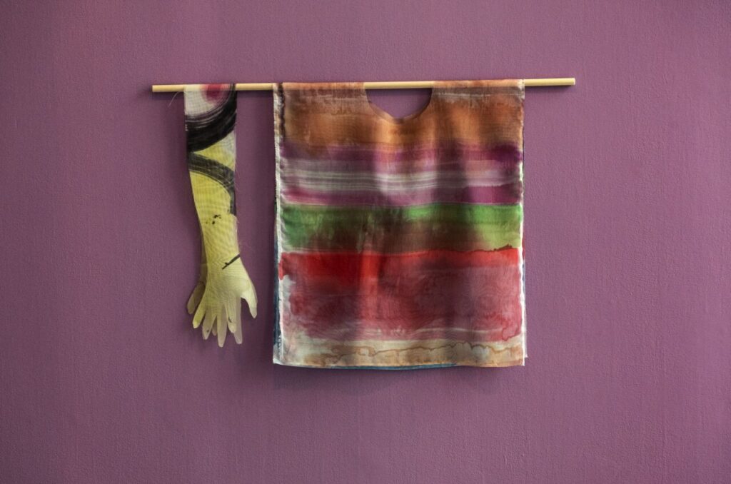 A hand-painted textile piece hangs on a purple wall. The artwork includes a colourful striped tunic-like form and a long sleeve shaped like an arm and hand, both suspended from a wooden rod.