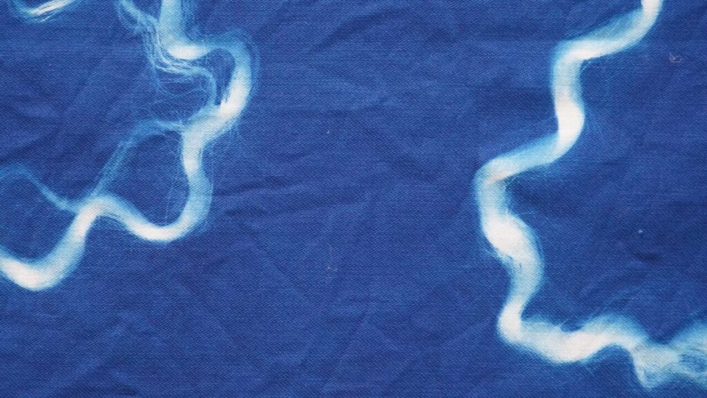 An abstract cyanotype print featuring soft white, wavy lines on deep blue fabric. The organic, fluid shapes evoke movement and energy against the textured surface.