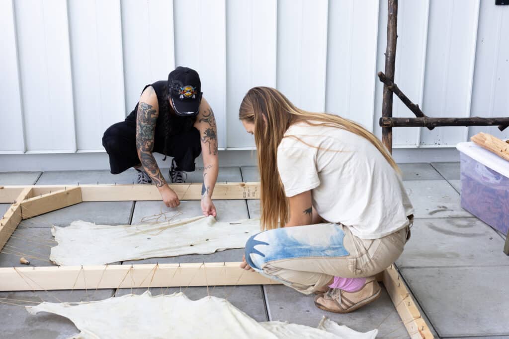 Two people crouch on the ground, stretching and tying rawhide to a wooden frame as part of a hide tanning workshop.