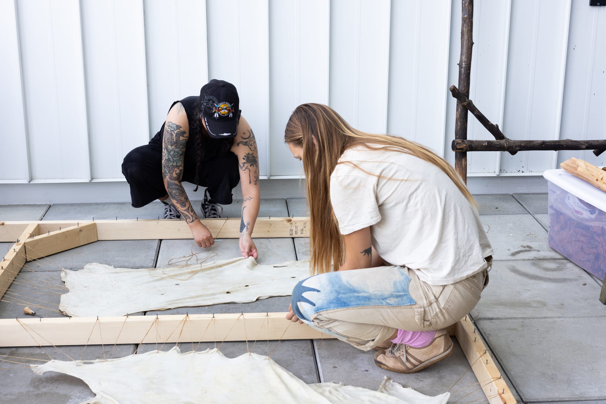 Two people crouch on the ground, stretching and tying rawhide to a wooden frame as part of a hide tanning workshop.