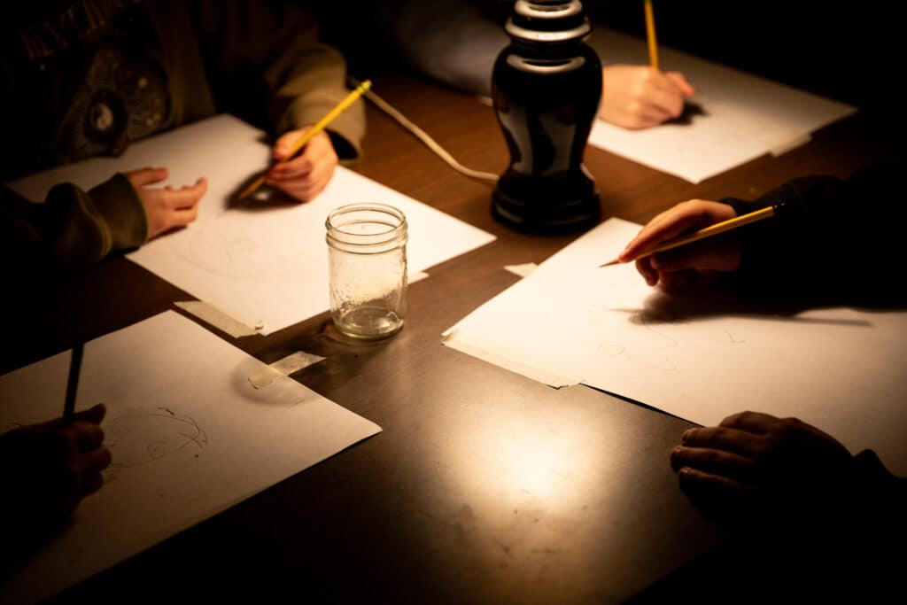 Hands draw with pencils on sheets of paper lit by a single lamp on a table.