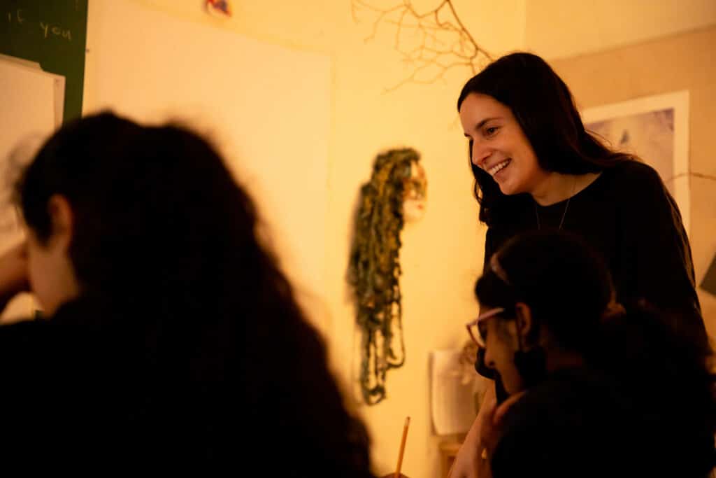 An instructor smiles while engaging with students in a warmly lit classroom.