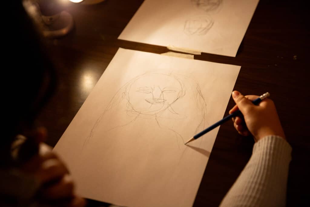 A person sketches a portrait in pencil on white paper under warm lamplight.