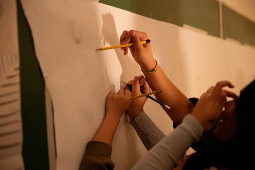 Hands holding pencils draw together on a large sheet of paper taped to a wall.