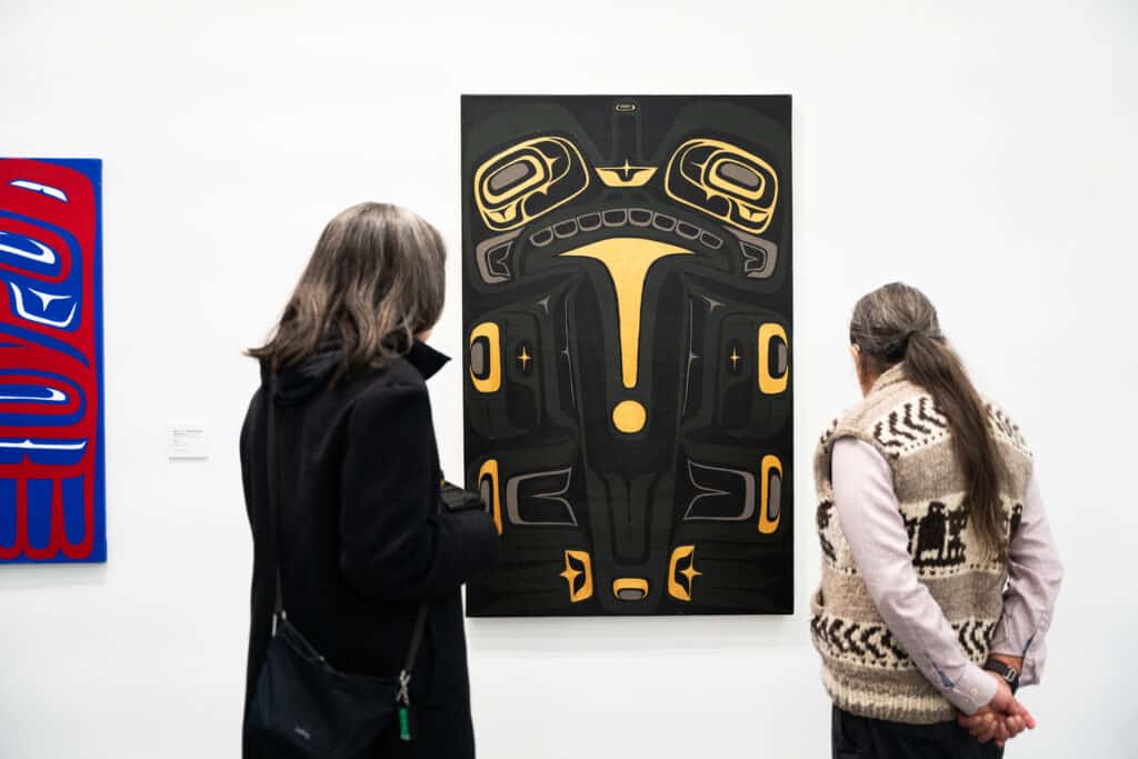 Two people view an Indigenous artwork in a gallery. The painting in front of them features a symmetrical black and gold design in a Northwest Coast style, displayed on a white wall. One person wears a dark coat and crossbody bag, while the other wears a patterned wool vest and has long hair tied back.
