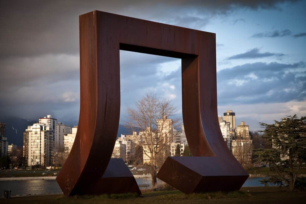A large rust-coloured steel sculpture shaped like an oversized staple stands on a grassy area near False Creek with downtown Vancouver in the background.