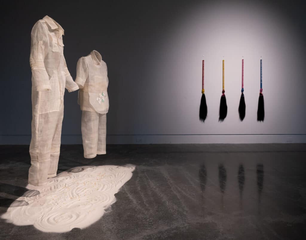 A gallery installation showing two translucent traditional garments made from natural fibre fabric standing over a bed of white sand, with four hair brooms wrapped in coloured bands hanging on the wall behind them.