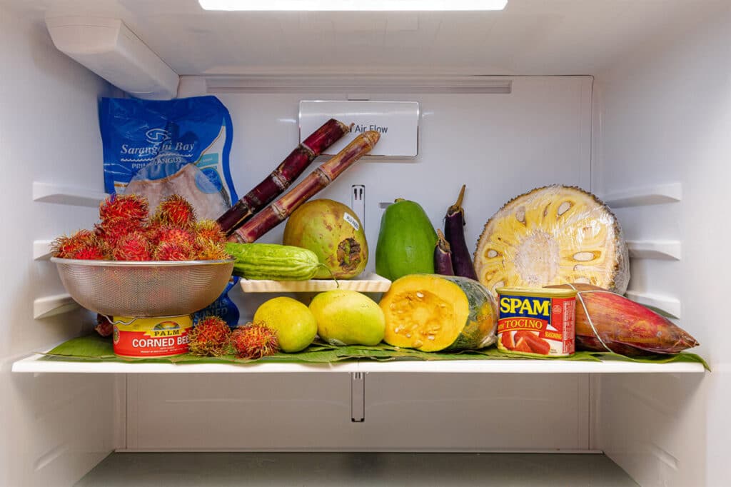 Inside view of a refrigerator filled with tropical and Filipino foods, including rambutan, jackfruit, bitter melon, mangoes, sugar cane, canned corned beef, Spam Tocino, and frozen fish, arranged neatly on banana leaves.