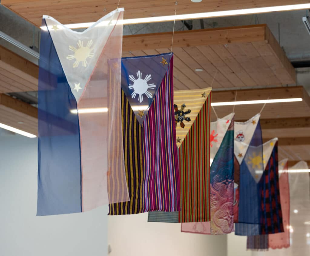 A series of textile artworks hang from the ceiling, inspired by the design of the Philippine flag, each rendered in different fabrics, colours, and patterns.