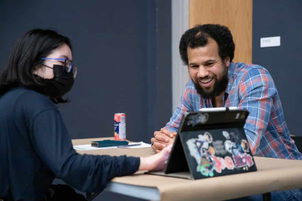 A student and an advisor sit at a table having a conversation, both smiling while looking at a laptop covered in stickers.