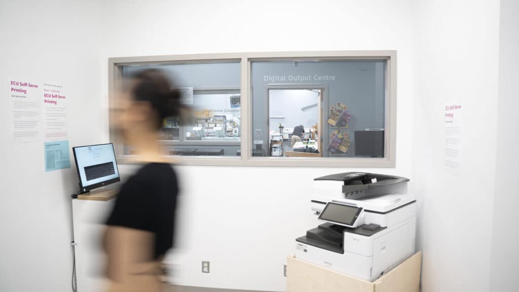 Large-format Canon printers lined up beneath windows that look into the Digital Output Centre, with signs for self-serve and output computers.
