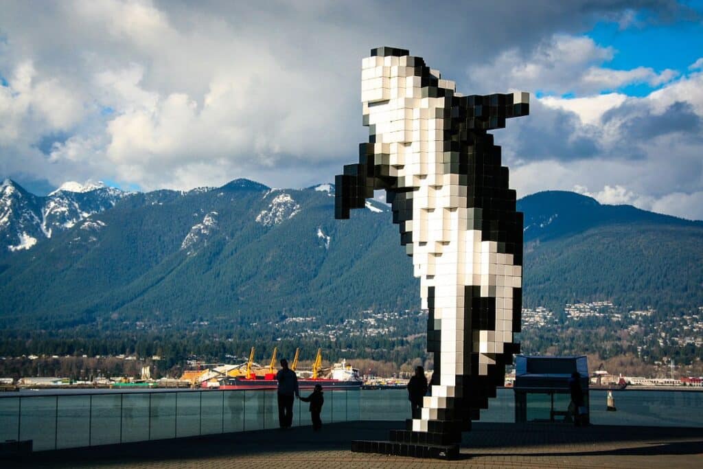 A large black-and-white sculpture shaped like a pixelated orca whale stands near Vancouver’s waterfront with mountains in the background.