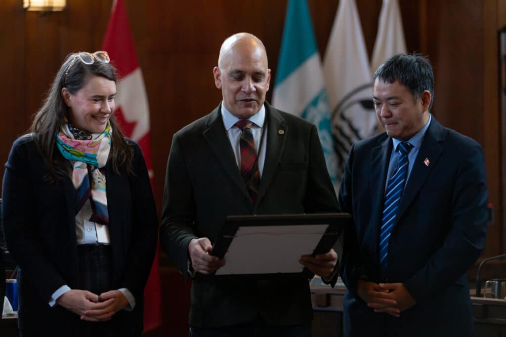Three people stand together in Vancouver City Hall, one holding a framed document while reading from it.