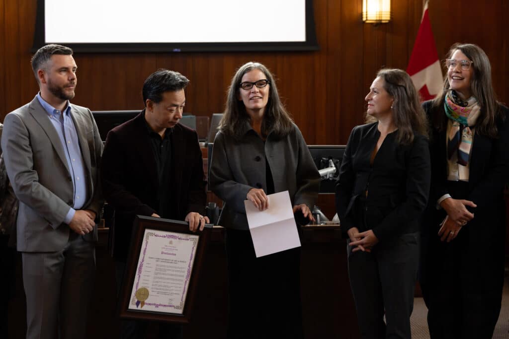 Five people stand together in Vancouver City Hall, holding a framed proclamation and a printed speech.