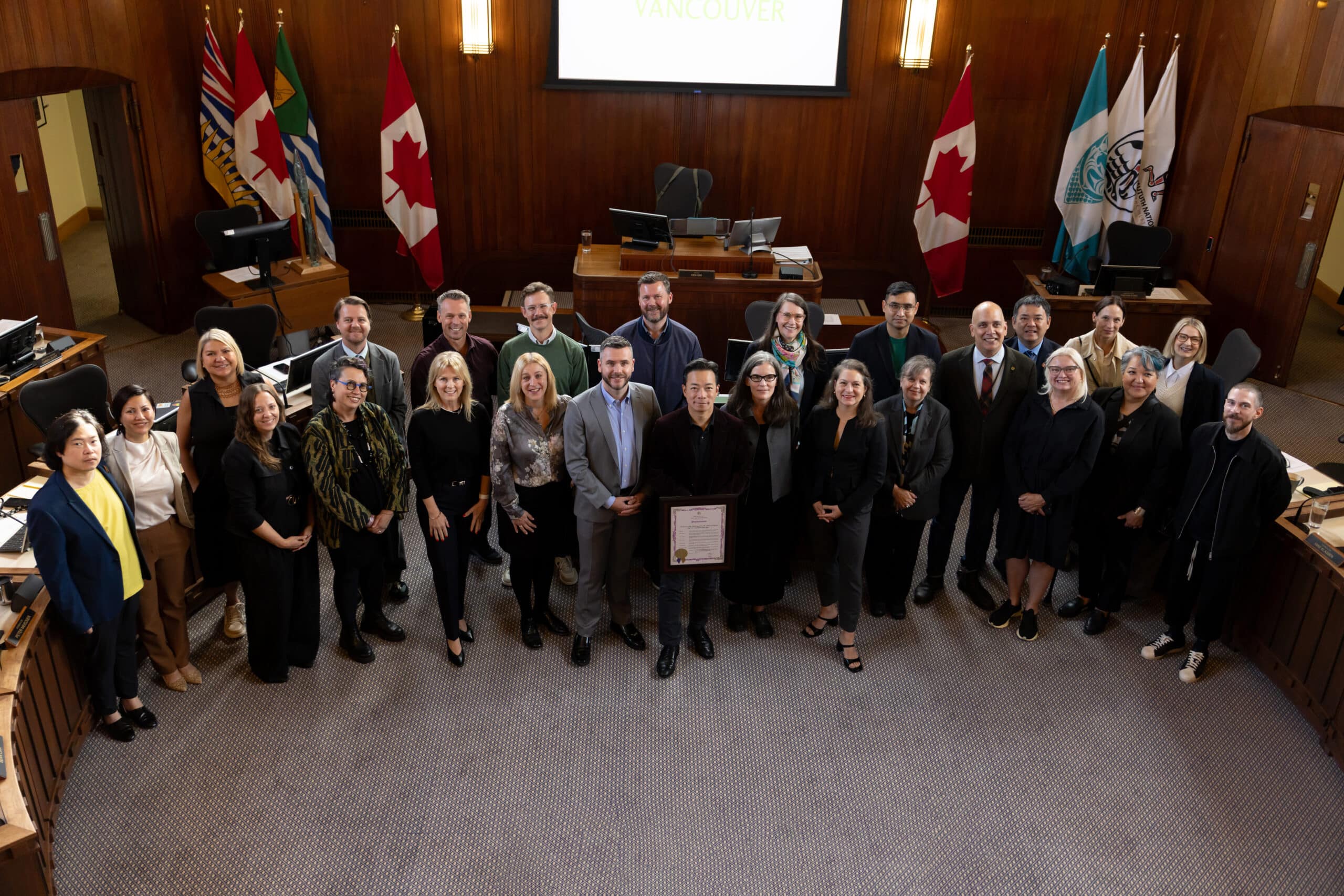 A large group of people stands together in Vancouver City Hall chambers for a group photo, smiling at the camera.