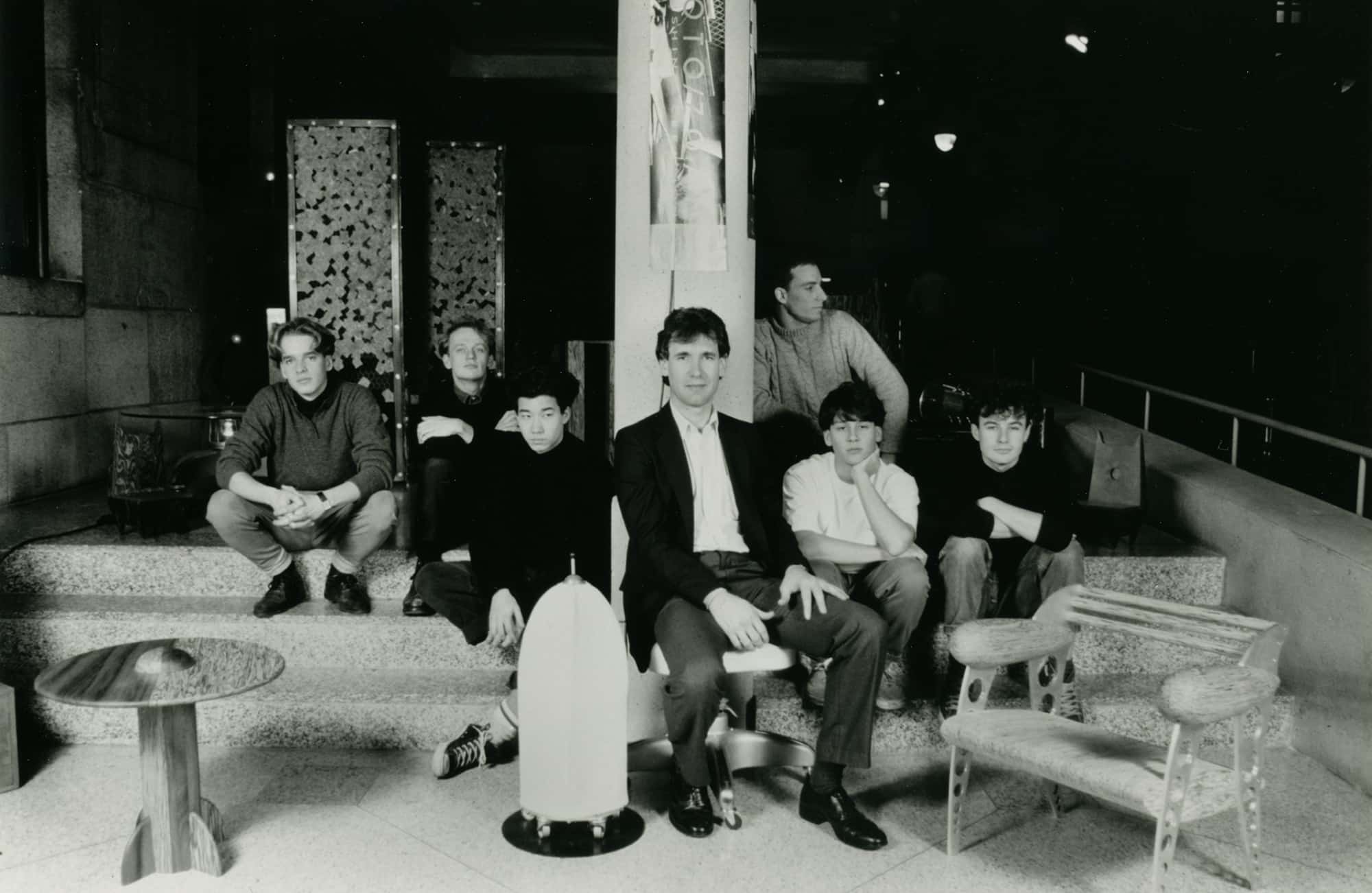 A group of seven men sit and pose on the steps of an art gallery surrounded by sculptural furniture and prototypes. The central figure in a suit sits among students in casual sweaters and jeans.