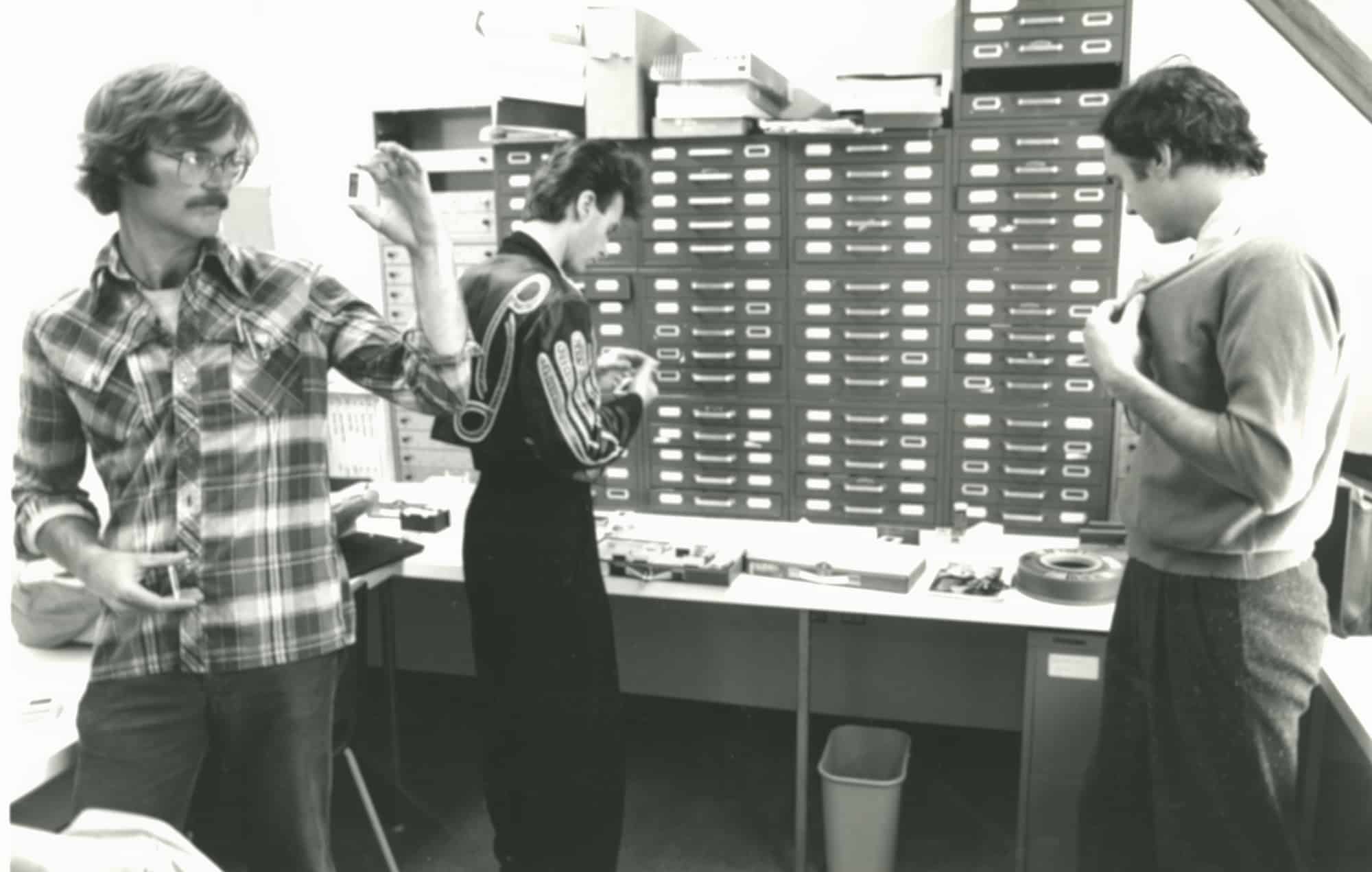 Three men in a design studio examining small mechanical parts. One man in glasses and a plaid shirt holds up a piece, while another in a patterned jacket looks down at his hands. Rows of labeled drawers line the back wall.