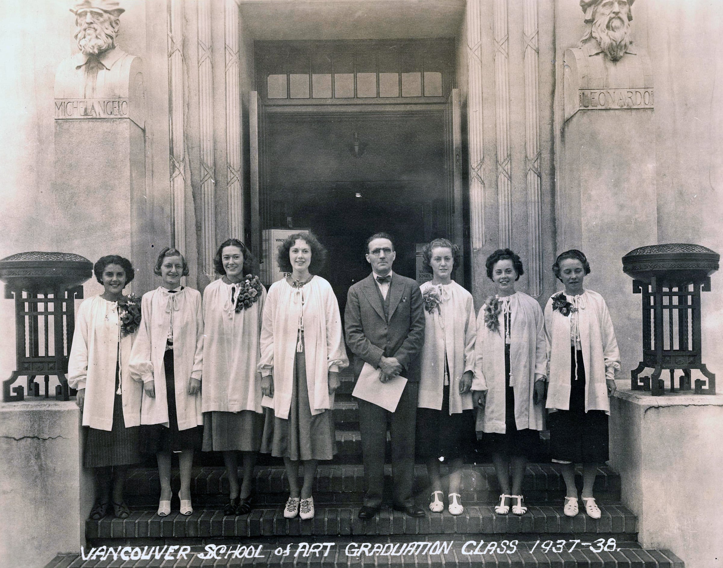 A black-and-white photo of the 1937–38 Vancouver School of Art graduation class. Seven women in light coats and floral corsages stand beside a man in a suit on the school steps. Sculpted busts of Michelangelo and Leonardo flank the entrance above them.