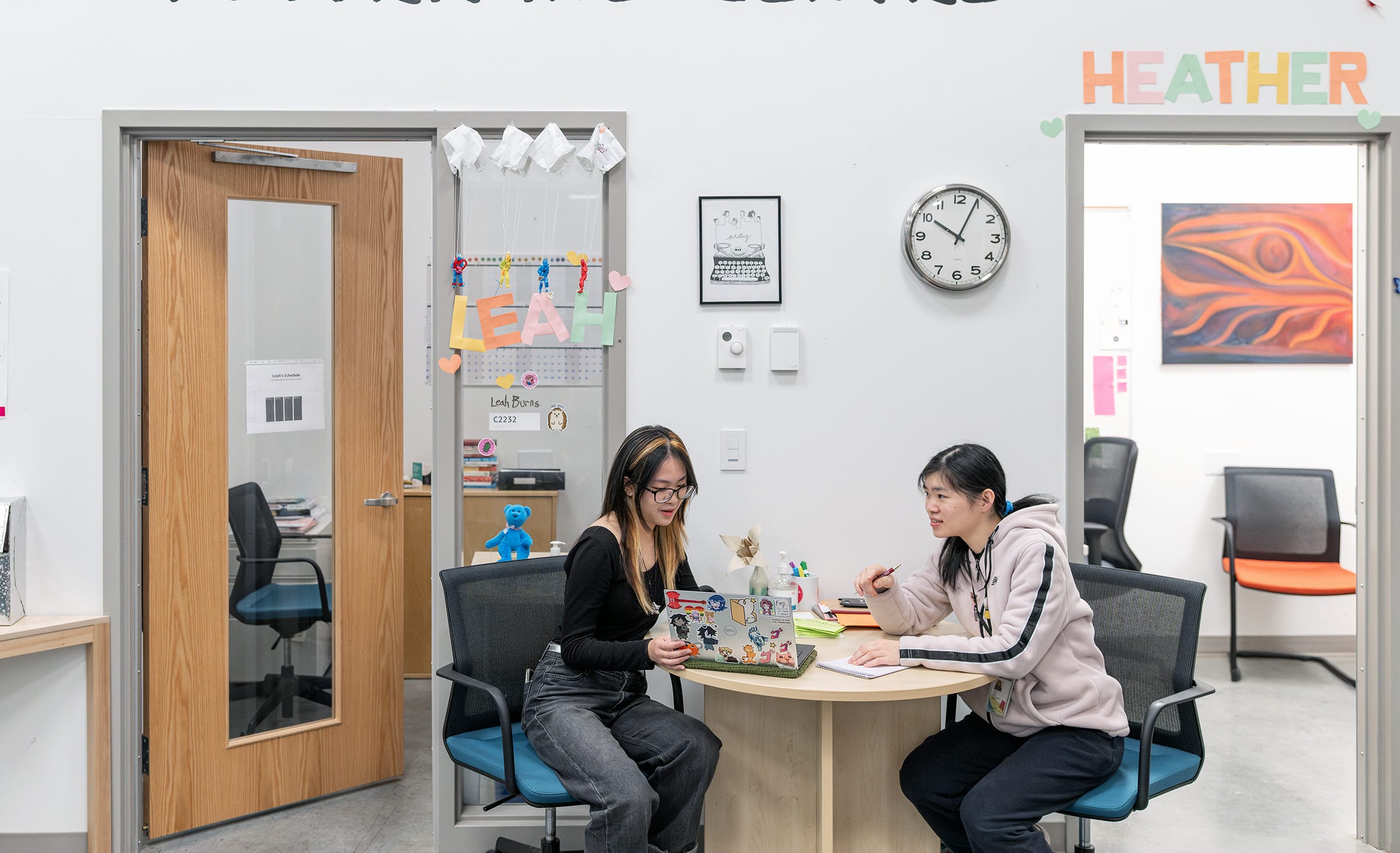 Two students sit at a small round table in a bright office space, working together. One student with glasses uses a laptop covered in stickers, while the other writes in a notebook. Behind them are two office doors decorated with colourful cut-out names, “LEAH” and “HEATHER.” A clock and artwork hang on the wall, and office chairs are visible in the adjoining rooms.