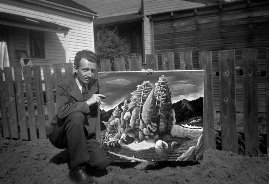 A man kneels in a yard, pointing to one of his landscape paintings displayed against a fence.