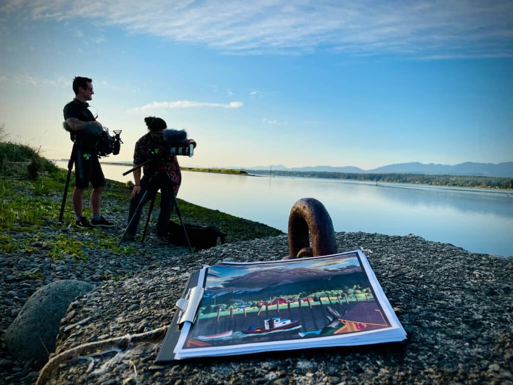A film crew sets up cameras beside a calm shoreline, with a painting on a rock in the foreground.