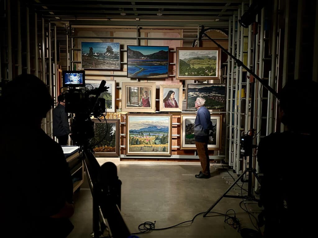 A man views framed paintings in a gallery storage area, surrounded by camera equipment.