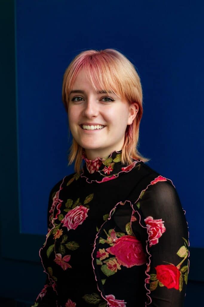A person with short, light hair smiles while wearing a black floral top against a deep blue background.