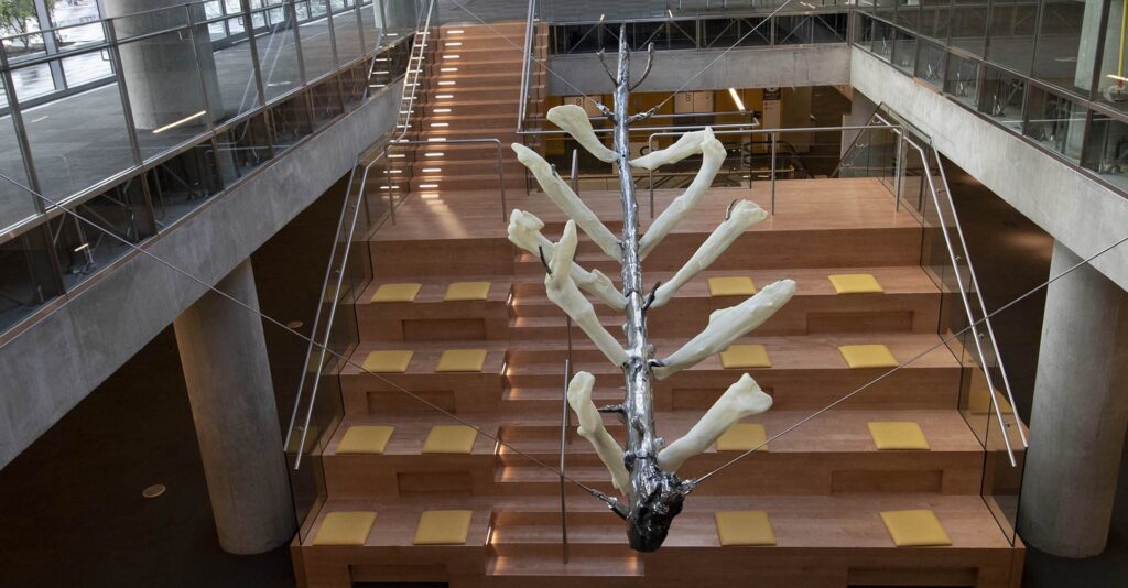 A metallic and resin sculpture resembling a tree branch hangs in the atrium of a modern university building.