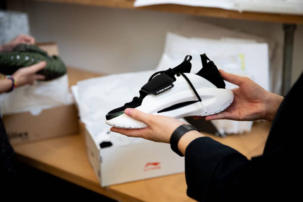 Close-up of a person holding a black and white athletic shoe over an open shoebox on a table, with another person holding a green shoe in the background.