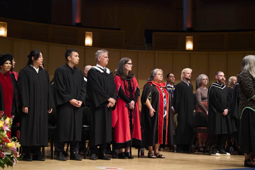 Faculty and graduates stand on stage in their regalia during the convocation ceremony. The setting is a concert hall with warm lighting and a wooden backdrop.