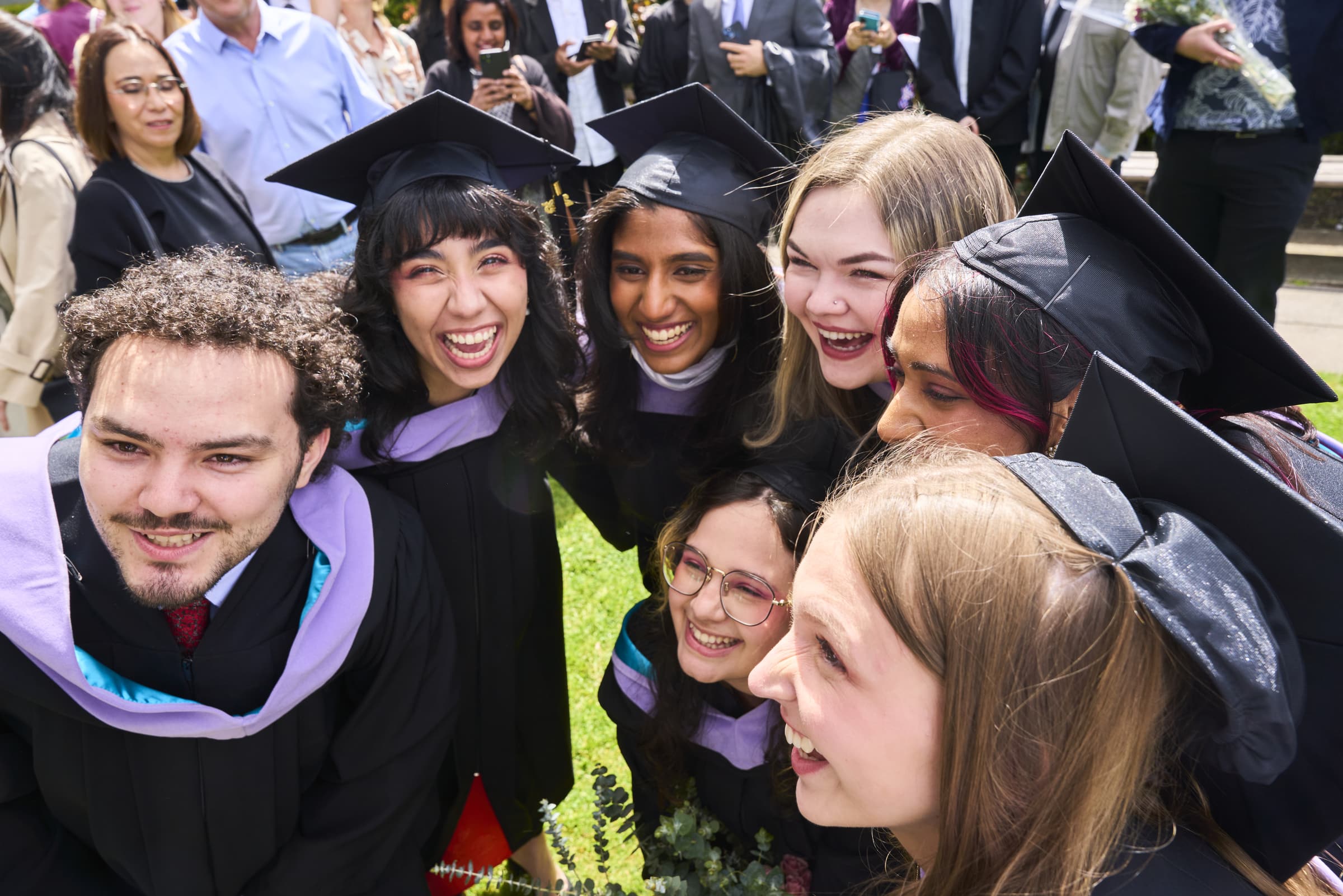 A group of ECU graduates in black gowns and purple hoods lean together, laughing and smiling for a group photo surrounded by friends and family at convocation.
