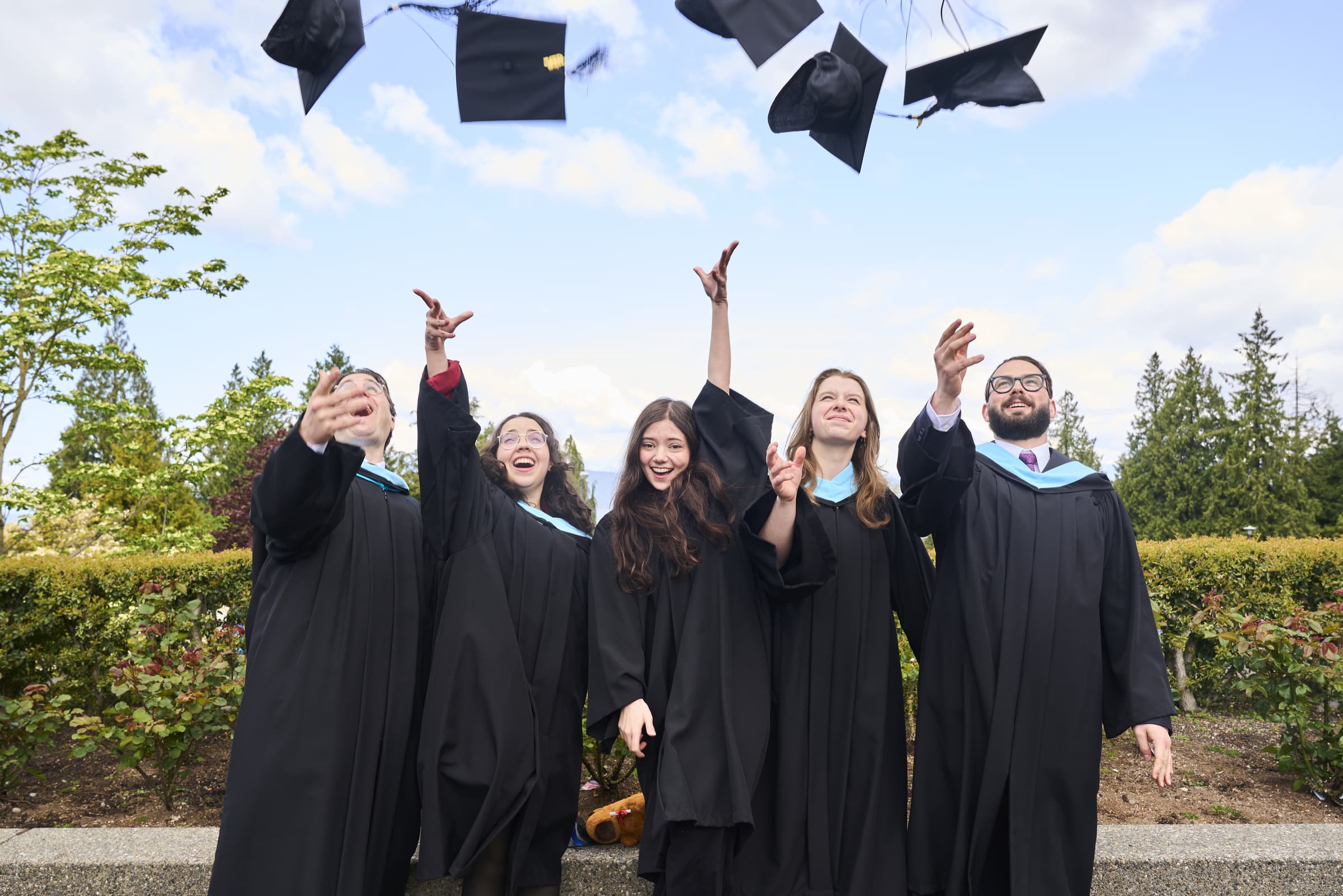 Five ECU graduates in black gowns and blue hoods celebrate by throwing their caps in the air outdoors against a bright, partly cloudy sky.