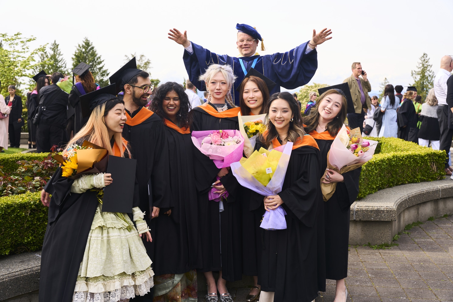 A group of ECU graduates in black gowns and orange hoods stand outdoors, smiling and holding bouquets. Behind them, a faculty member in blue regalia raises his arms in celebration during convocation.