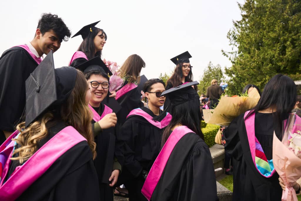 A group of ECU graduates wearing black gowns with pink hoods gather outdoors after convocation, laughing and chatting while holding bouquets of flowers.