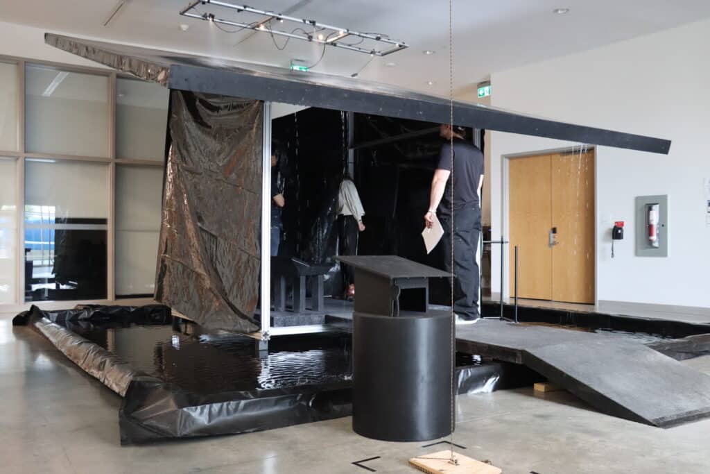 An installation featuring a dark, angular structure made of black panels and tarps stands in the middle of a gallery space, surrounded by shallow water contained in a plastic barrier. A ramp leads into the structure where a few people explore the dimly lit interior, giving the scene an immersive and atmospheric quality.