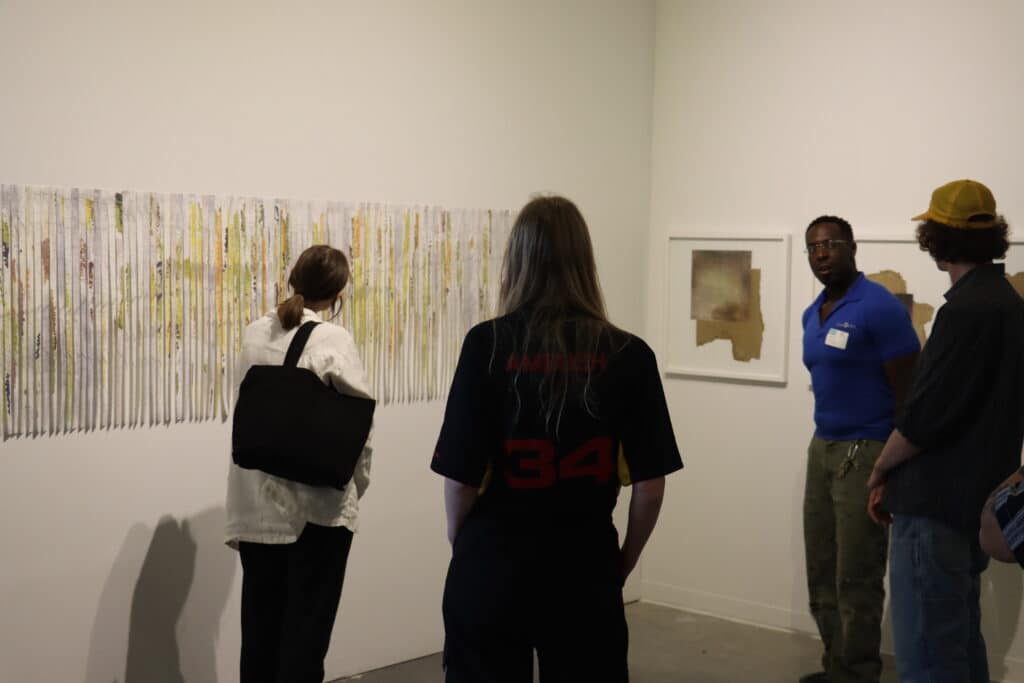 A small group of people stand in a gallery viewing a long, textured artwork made of vertical strips of paper or fabric with soft washes of yellow, green, and brown. Two framed abstract pieces hang nearby, and one person speaks while the others listen attentively, suggesting an artist-led discussion.