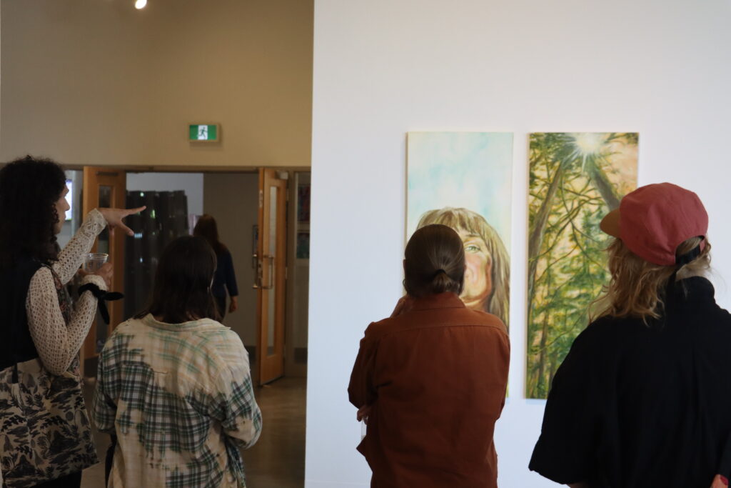 A small group of people stand in a gallery viewing two vertical paintings side by side—one depicting a person’s upward gaze, the other showing tall trees with sunlight filtering through. One person gestures animatedly while holding a drink, creating a sense of lively discussion and shared reflection.