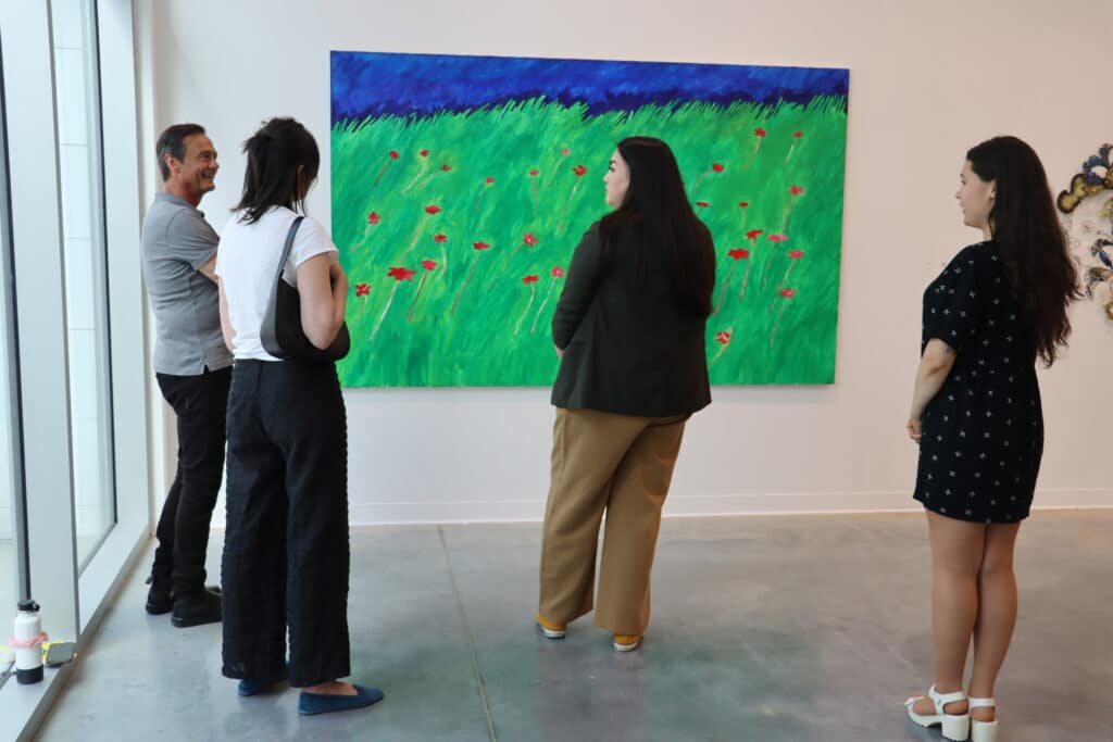 Four people stand in a bright gallery space viewing a large painting of a green field dotted with red flowers beneath a deep blue sky. The group appears relaxed and conversational, with sunlight streaming through nearby windows.