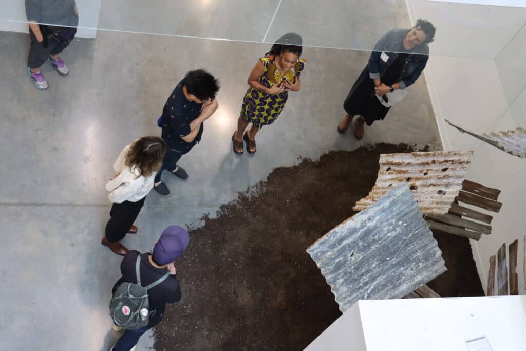 Viewed from above, a small group of people gathers around an installation made of rusted corrugated metal sheets arranged over a patch of soil. The group stands in conversation, observing the work within a spacious gallery setting.