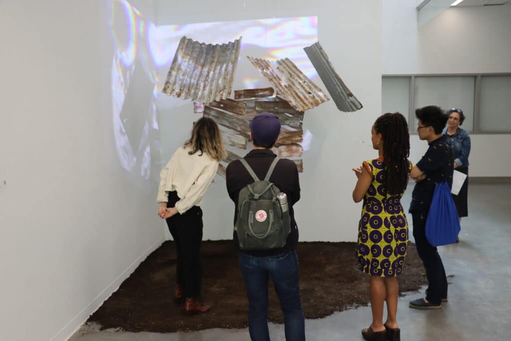 A small group of people observe an installation made of rusted corrugated metal sheets arranged against a white wall, with projected light and shadows creating shifting reflections. The piece sits on a patch of soil, blending natural and industrial textures within the gallery space.