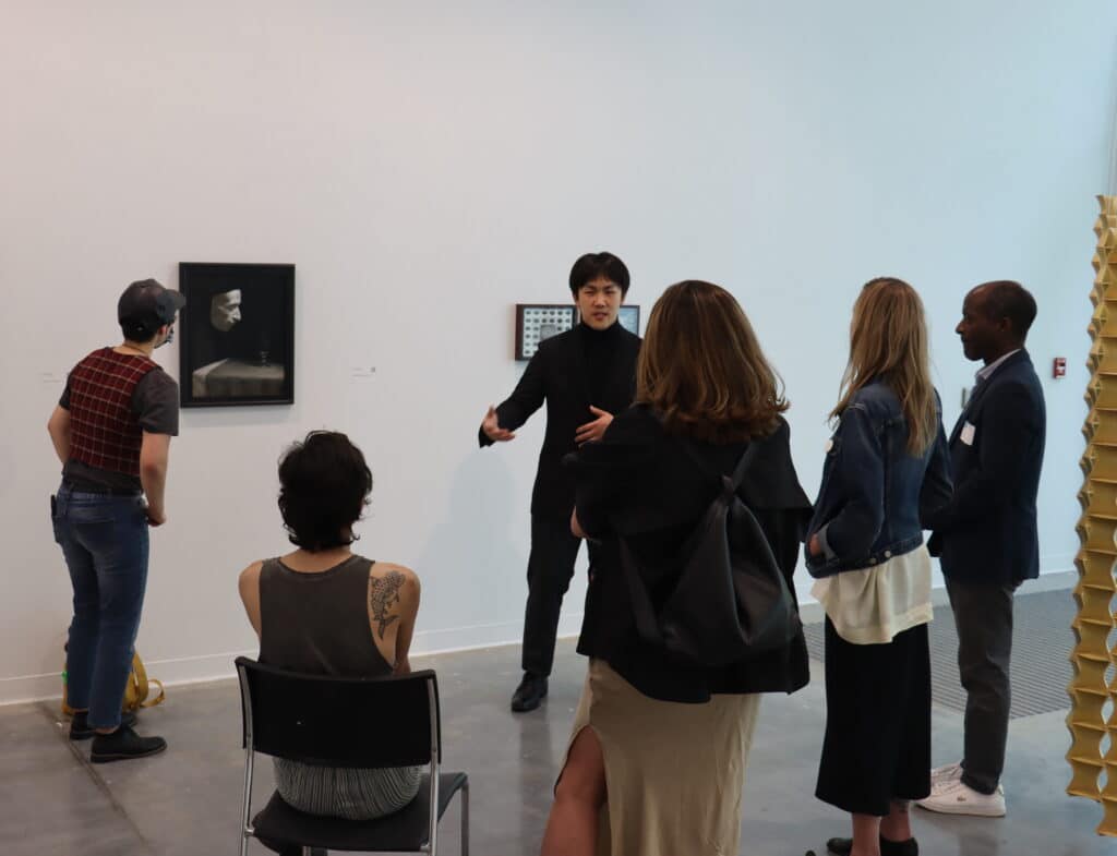 A person dressed in black speaks to a small group gathered in an art gallery. The group stands in front of two framed artworks on a white wall, engaging in conversation. The atmosphere appears lively and attentive, suggesting a guided tour or artist talk in progress.