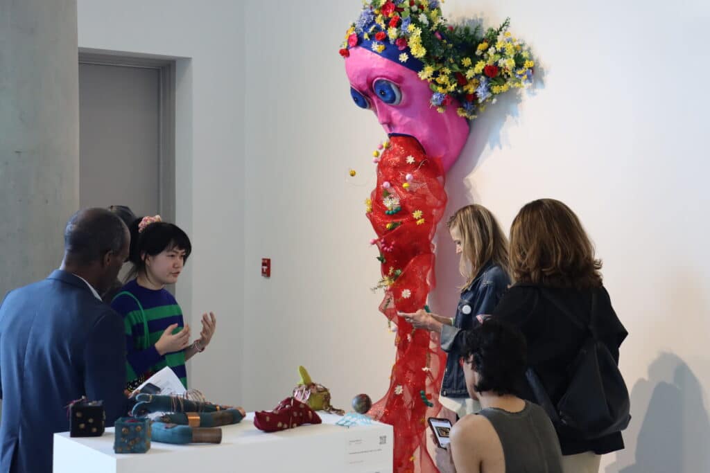 A group of people gather around a colourful wall sculpture of a large pink face with wide blue eyes and a cascade of flowers for hair. A red mesh material decorated with small flowers and objects flows from the mouth, creating a dramatic, whimsical effect. One person gestures while speaking, suggesting an animated discussion about the artwork.