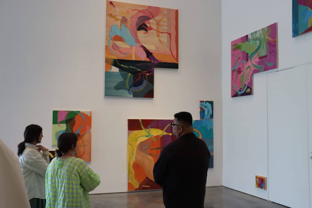 People stand in a gallery space observing a vibrant series of abstract paintings. The works feature sweeping brushstrokes and bold colours in shades of orange, pink, teal, and yellow, arranged asymmetrically across a white wall. The scene captures a quiet moment of reflection and discussion.