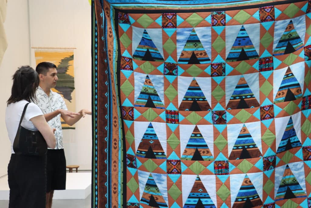 A person gestures while speaking to another viewer beside a large, colourful quilt featuring geometric patterns and triangular shapes resembling tipis. The fabric is composed of earthy tones mixed with bright blues and greens, blending traditional motifs with contemporary design.