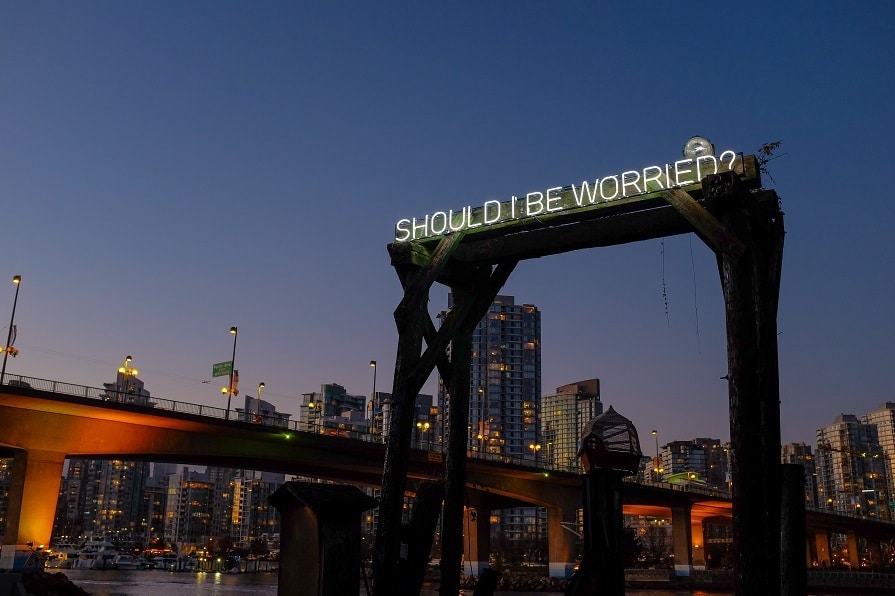A glowing neon sign reading “Should I Be Worried?” is mounted on wooden beams near the water, with city buildings illuminated at dusk.