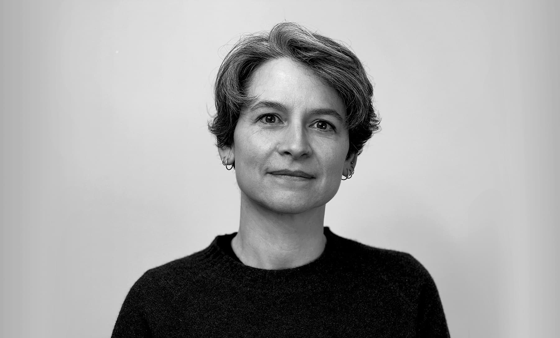A black-and-white portrait of a person with short hair wearing small hoop earrings and a dark sweater, looking calmly toward the camera against a plain background.